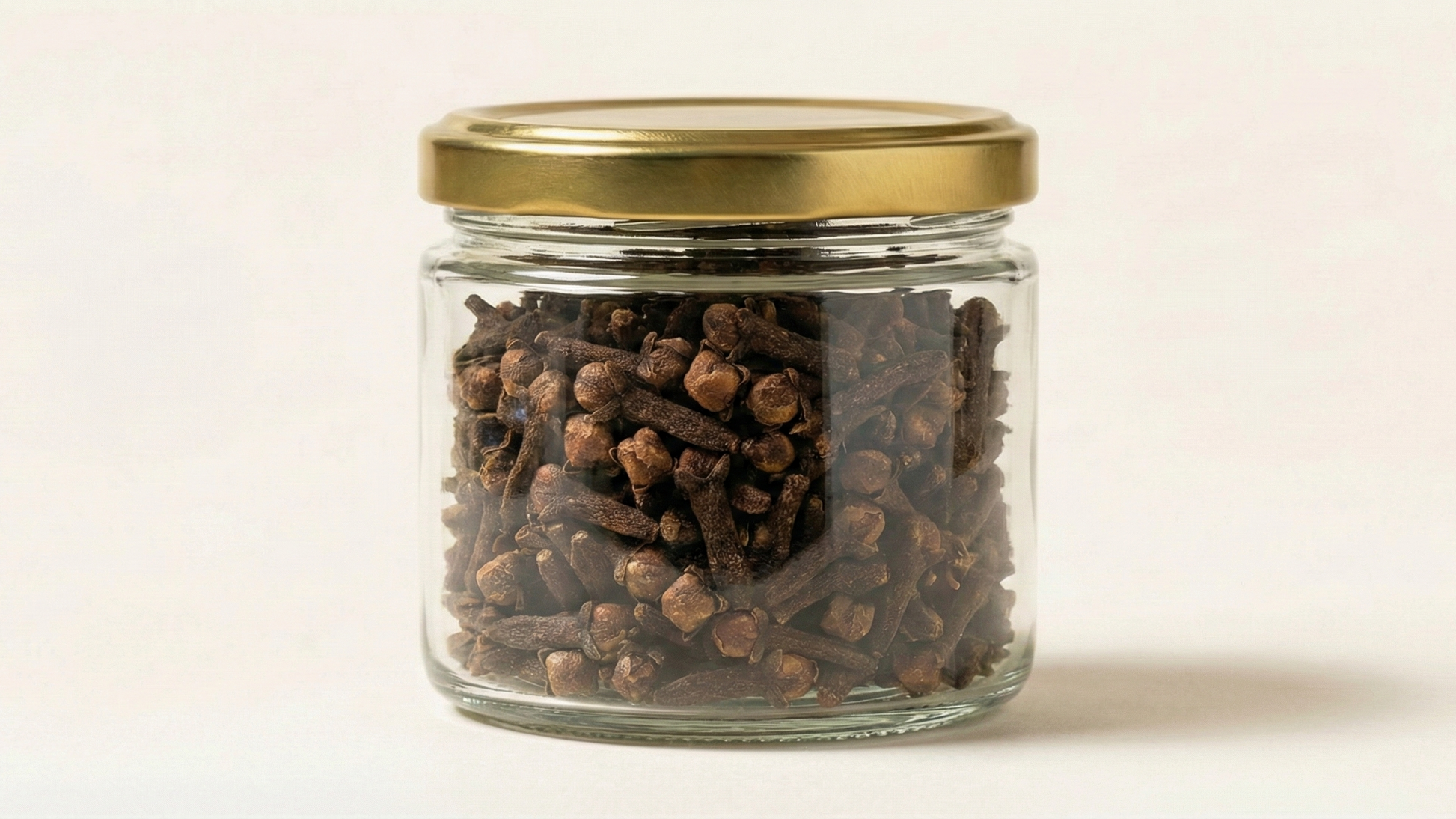 Glass jar with gold lid filled with cloves on a light beige background